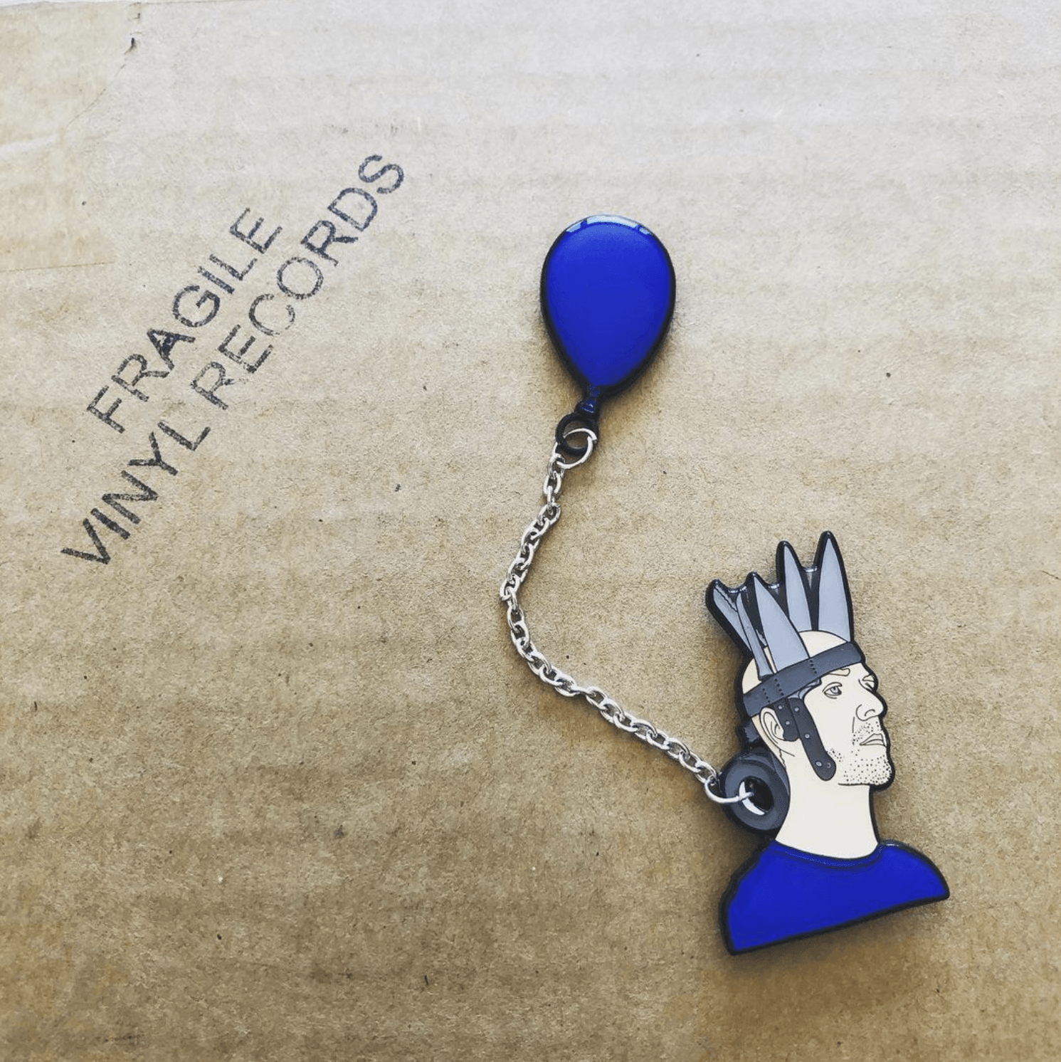 jan hakon erichson enamel pin with Jan wearing a red shirt and crown of duct taped knives with a blue balloon attatched to a chain goin through the duct tape roll. the pin is stuck into a piece of cardboard the reads "fragile, vinyl records."