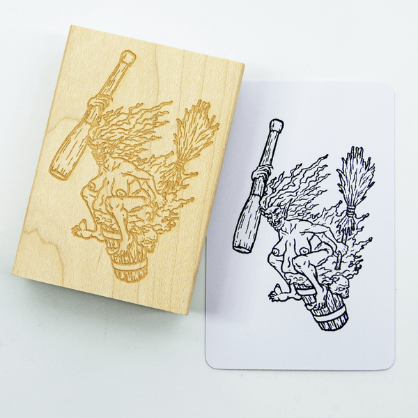 A wood and rubber stamp of Baba Yaga the crone witch, riding her mortar and pestle with broom in hand. She has taken off her clothing and sits on it with a wicked smile. A stamped white notecard shows the design in black ink.
