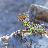 A green, yellow, black, orange, blue, and gold enamel pin of the Citheronia Regalis caterpillar, the Hickory Honrned Devil, placed upon a log and sparkling in the daylight. 