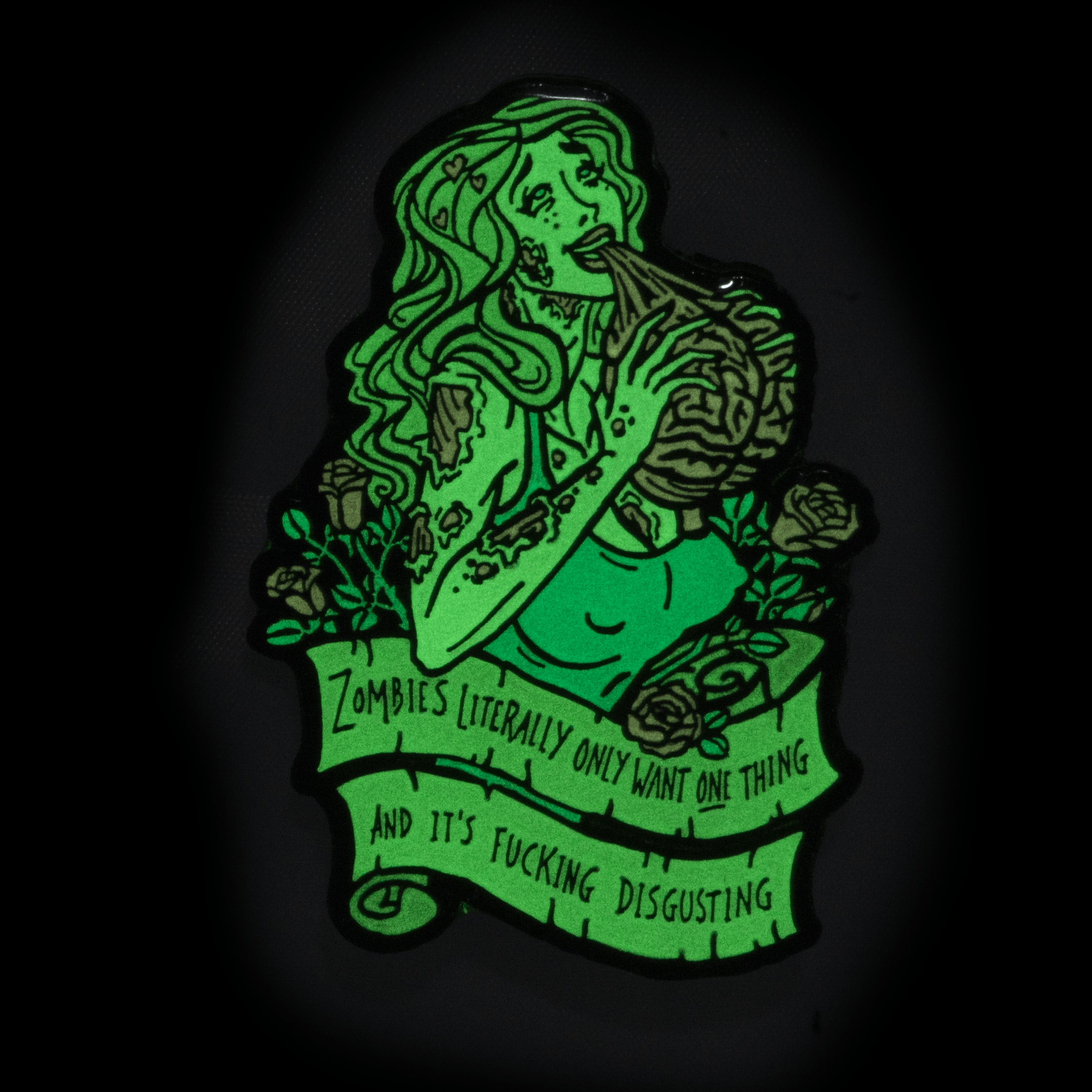 An enamel pin of a zombie woman eating a brain. An er below her says "Zombies literally only want one thing and it's fucking disgusting". The pin is glowing green in the dark.
