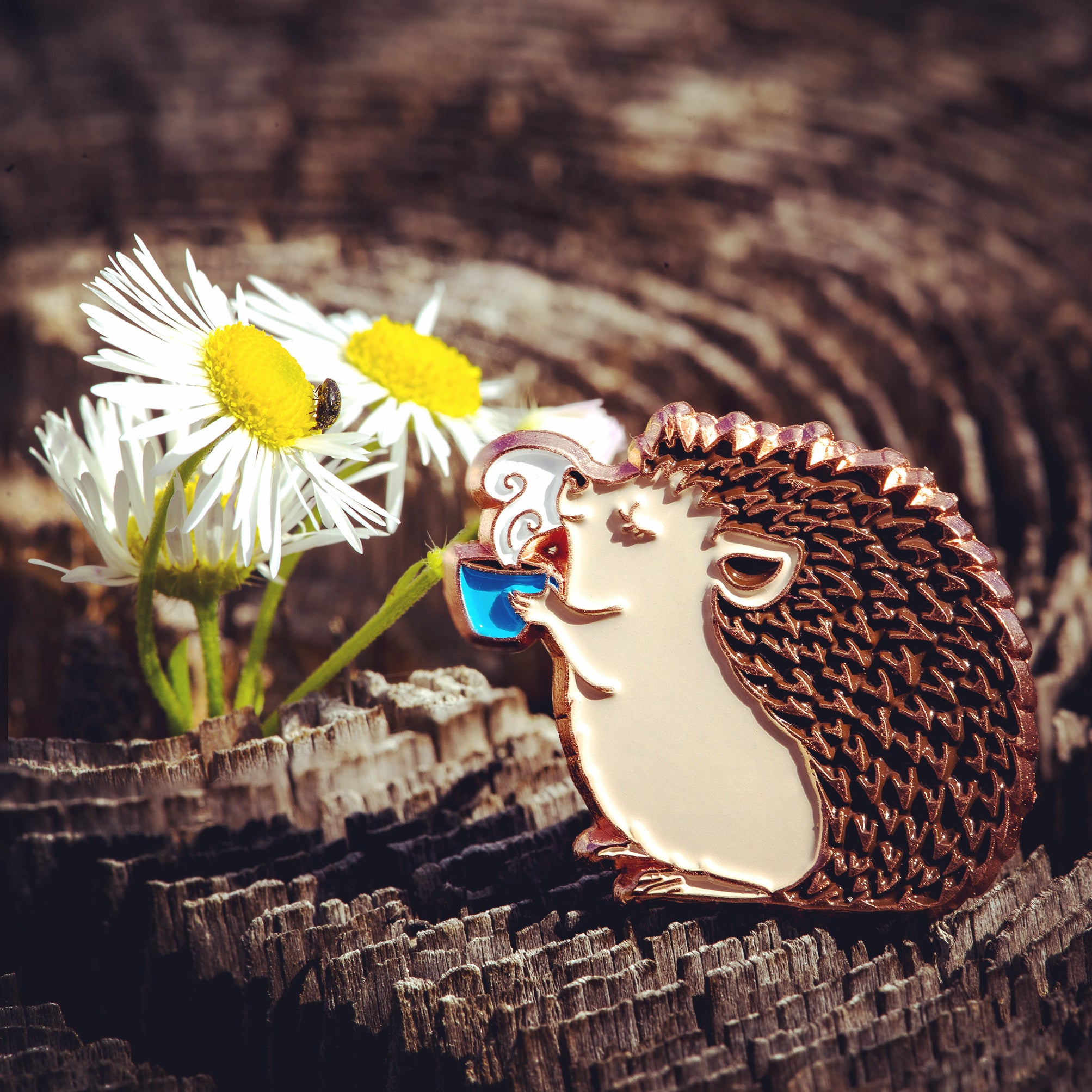 An enamel pin of a hedgehog enjoying a steaming blue mug of coffee, next to some daisies and a beetle.