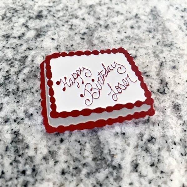 An enamel pin in the shape of a white and red birthday cake, with faux icing text that says "Happy Birthday Loser". This image was featured in many viral articles.