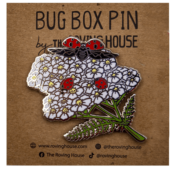 An enamel pin of three red and black nine-spotted ladybugs exploring white yarrow blossoms.