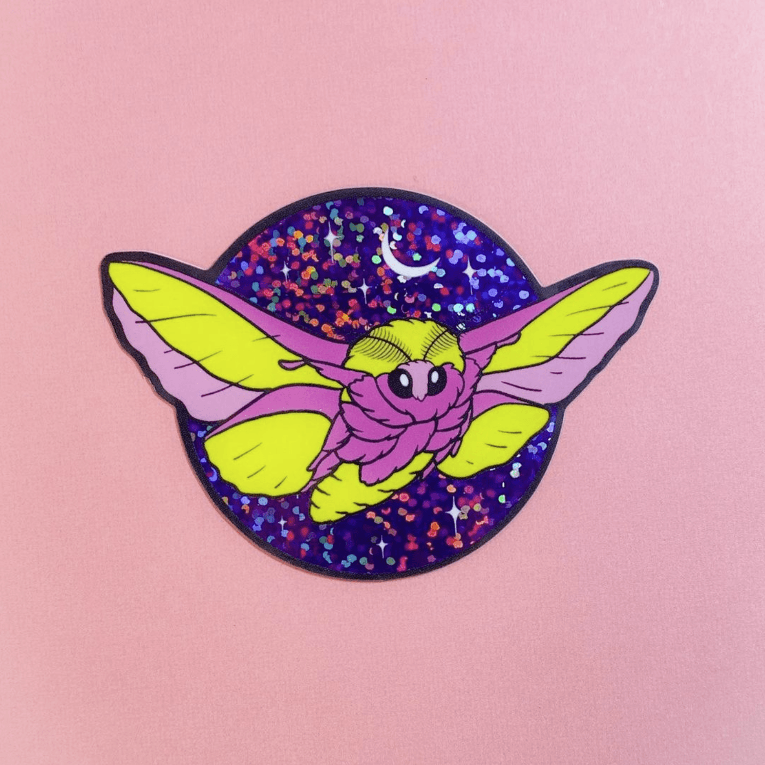 A glittery sticker of a pink and yellow rosy maple moth with big black eyes, flying in the starry night sky with the moon overhead.