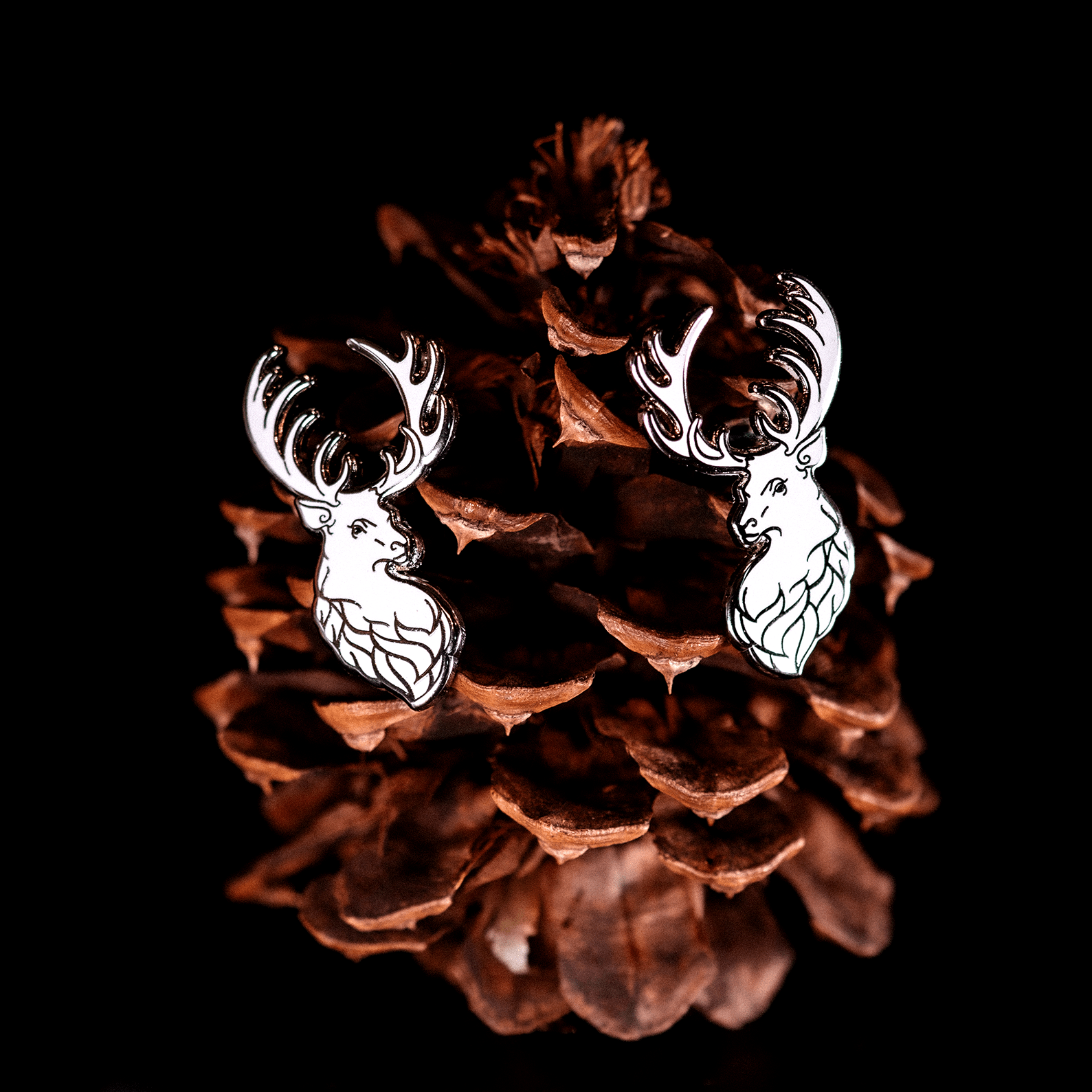 A pair of white winter stag pins by The Roving House, positioned on a pinecone.